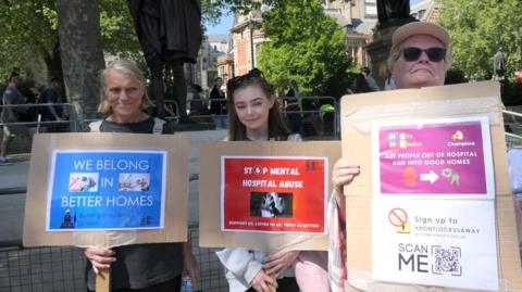 Three woman pose for a photo holding placards protesting about the detention of people with autism. The two either side are around 60 years of age while Lucy, in the centre, is in her twenties. Lucy has long brown hair with sunglasses on her head. 