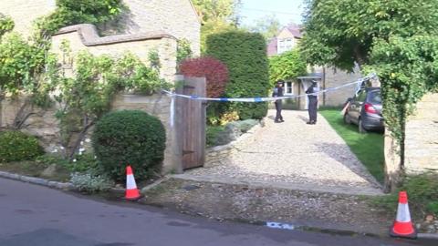 A stone driveway off a road with cones in place at the entrance - it leads to a house where two police officers are standing near the front door.