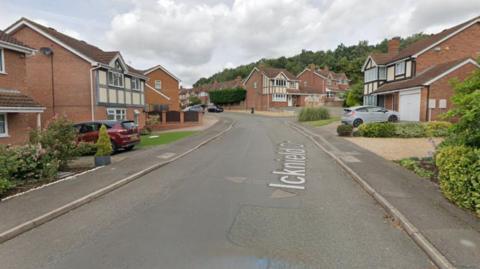 A street view of Icknield Drive in Northampton where the new care home is to be built. A grey tarmac road has detached red brick houses all along the road. Several cars are parked on the drives. 