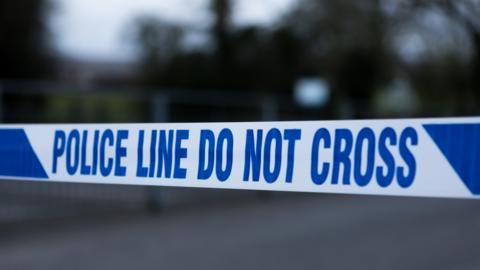 A line of blue and white police incident tape, with a blurred rural background.