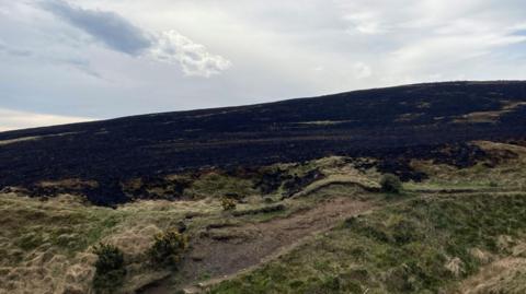 A wide stretch of moorland scarred by fire, with a large area of blackened, scorched ground running across a hillside.