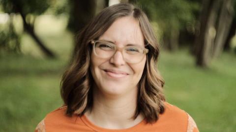 A smiling Clare Smyrell with mid-length light brown hair, glasses, wearing an orange top, with grass and trees seen blurred in the background