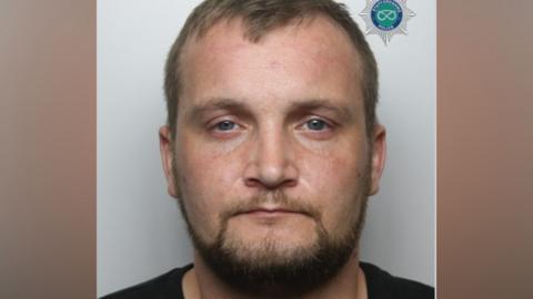 A police mug shot of a man with short brown hair and a beard. He is positioned against a light-coloured background and is looking directly at the camera. He is wearing a black top.