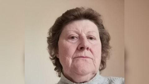 Head and shoulders shot of a woman with short curly brown hair looking into the camera wearing a grey jumper.