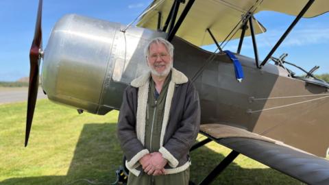 Tim Rayner, a white-haired man with a beard and glasses, stands in front of the plane, wearing a khaki flightsuit and a leather and sheepskin flying jacket. The plane's propeller is on the left of the picture.