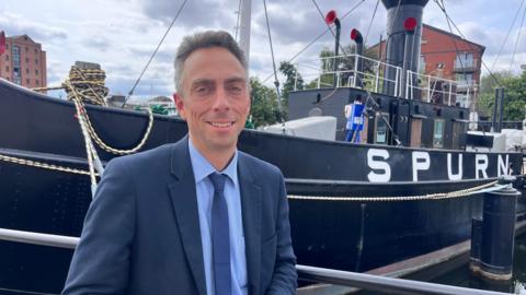 Mike Ross, the leader of Hull City Council stands in front of the Spurn Lightship which is berthed in Hull marina and looks directly at the camera wearing a navy blue suit and tie.