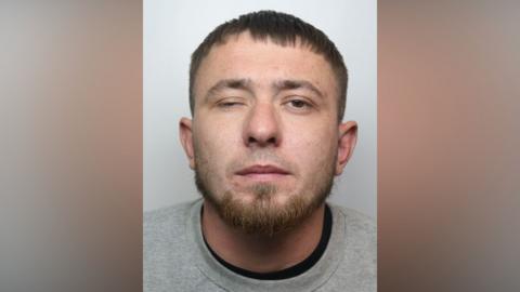 A police mugshot of a man with brown hair and a beard. He is wearing a grey jumper and is standing against a light-coloured backdrop.