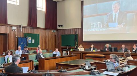 An older man wearing a suit and tie speaks to members of Cheltenham Borough Council in the chamber. He is reading off a sheet of paper and is projected onto a big screen for people to see. Councillors are looking at him.