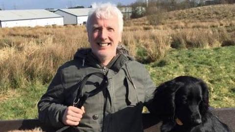 Brian Hurton sitting outside with his black dog, smiling at the camera 