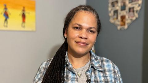 Nichola Howells is a lady in her late 40s, she is of mixed heritage and has long black dreadlocks in a ponytail to the side. She is wearing a chequered shirt and a necklace with a triangular pendant. She is looking directly at the camera in her living room. 