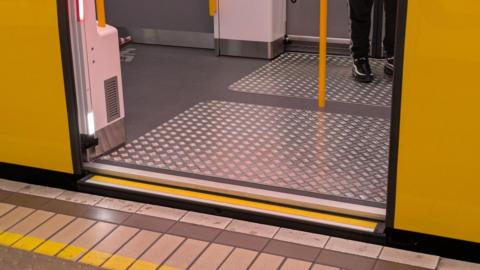 A train with its carriage door open, revealing a metal zig zag plate on the floor, with a person standing on it. The yellow doors are open and the platform is next to the open door. 