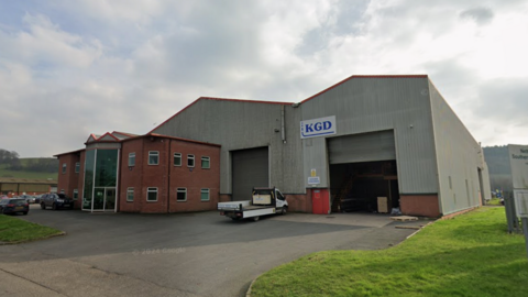 A large grey warehouse with red brick offices built on the front of it is pictured on a cloudy day. A flat bed truck is parked outside the main shutters to the warehouse.