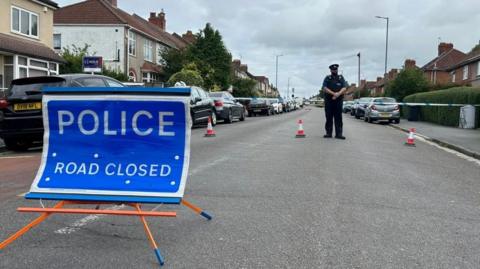 The police cordon in Bristol. There is a police road closed sign with a police officer and cones in the background