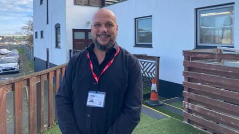 Justin Bellinger in a black shirt. He is wearing a red lanyard. Behind him is a white building and a car park. 