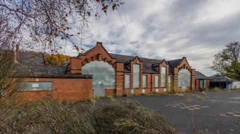 The old Victorian-era one-storey high Bluebell Meadow Primary School buildings in Trimdon Grange, County Durham. The school is boarded up with metal sheets, the playground is overgrown and unkept with weeds, while trees are blowing in the breeze on a grey day.