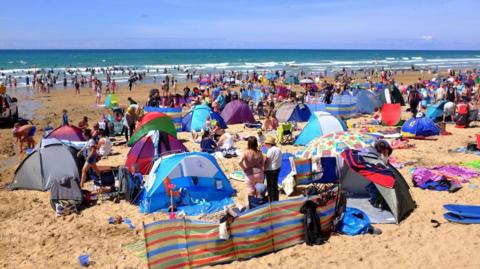 Dozens of people gathered on the beach at Watergate Bay in Newquay, Cornwall, during a sunny day. Several tents and beach wind screens have been set up on the sand. People are heading out into the sea on a sunny day.