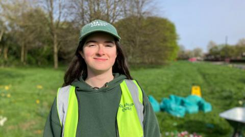 A girl with long black hair is smiling and looking at the camera. She is wearing a green hat and hoodie as well as a high-vis vest, all adorned with Cleanup Quest.