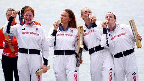 four women are stood on a platform wearing a white tracksuit with the red great Britain Olympic branding on their tops. They are all looking in random directions, Kissing their gold medals and holding a gold tube of a map in their opposite hands. 