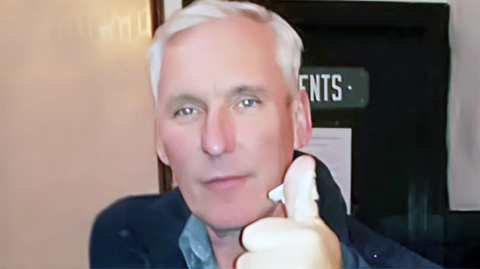 Jim Paterson is giving a thumbs up to the camera while standing outside a toilet door. He has short, white hair and is wearing a grey shirt and dark jacket.