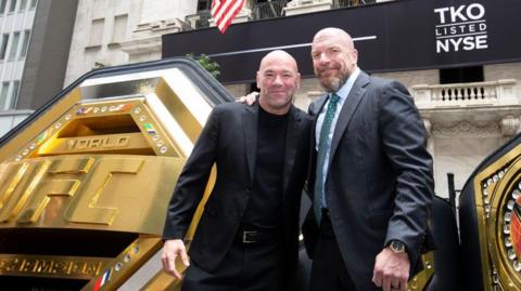 Dana White and Paul Levesque stand beside each other beside a massive UFC belt