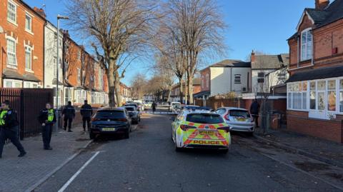 A residential street with parked cars on either side. There is police tape secioning off the road and a parked police car in the middle of the road just before the police tape.
