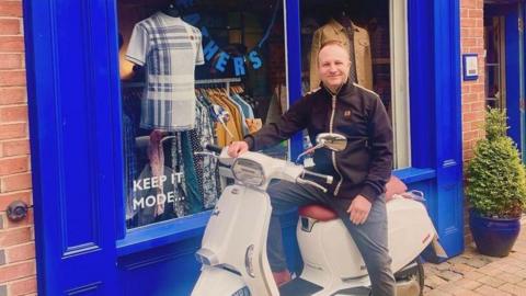 Man smiling while on a moped bike. he is wearing a black zip hoodie and dark blue jeans. he is sat outside a blue shop front