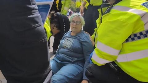 A woman in blue sweatshirt and jeans sits on the ground surrounded by police officers, who appear to be arresting her.