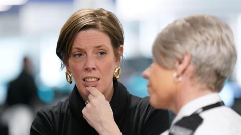 Jess Phillips wears a black jumper and gold earrings, her dark hair short, as she listens to a police officer with short grey hair and silver earrings.