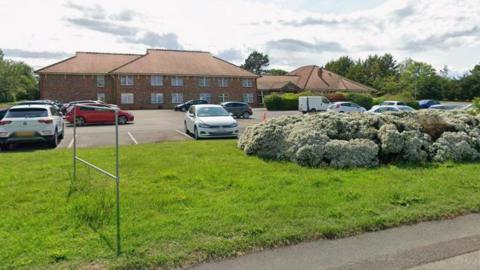 A hotel building with 14 windows visible, behind a car park which has about a dozen cars parked in it.