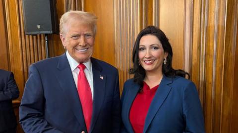 Donald Trump gives a thumbs up as he meets Deputy First Minister Emma Little-Pengelly. She is wearing a red dress with a blue blazer. The pair are standing in a wooden panelled room.