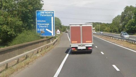 The A1(M) showing a blue and white sign for Stevenage (North), a lorry on the road, and two carriageways. There are trees around the road with cars on the opposite carriageway. 