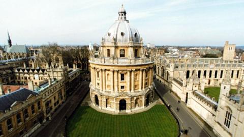 An aerial view of Bodleian Library with Oxford University colleges surrounding it. The lawn to the front of the library is visible as well. 
