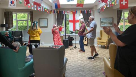 Several people are singing and dancing in the middle of a main room in a care home. There is a large England flag on the window in the back 