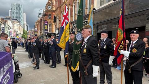 A ceremonial parade taking place on Briggate in Leeds. Shopfronts, street lighting, and tall structures stretch into the background. In the foreground a line of uniformed men stand dressed in dark outfits, some wearing berets or caps, and many have medals pinned to the front of their jackets. Some are wearing white gloves and holding tall ceremonial flags upright. 