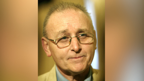 A close up image of Denis Donaldson. He has thinning, short grey hair and has a neutral expression on his face as he takes a question at a press conference. He has a pair of silver glasses on. He is wearing a tan suit jacket and a blue collared shirt.