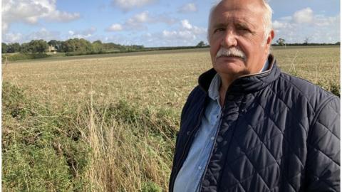 Man with white hair and moustache wearing a padded jacket stands in a field