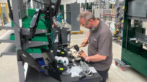 A man makes something at a workstation in a warehouse. He is wearing gloves and goggles.