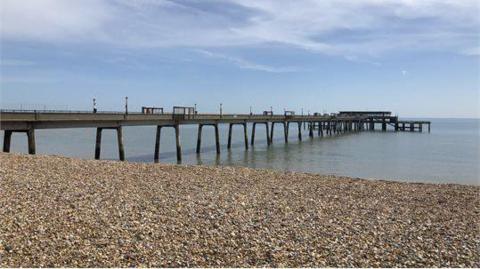 Deal pier reaches out into the sea, with the photo taken on a stony beach. The sea is calm and the sky is blue while the pier looks empty.