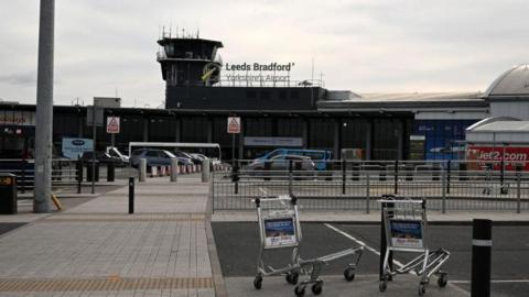 An airport building with a sign reading Leeds Bradford Yorkshire's Airport on the front. Rows of bollards and fences can be seen in front of the building. Two abandoned luggage trolleys are in front of the building.