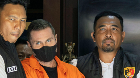 Steven Lyons, wearing a black face mask and an orange boiler suit, is flanked by police officers