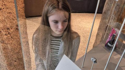 Bethan - a young girl sat by a glass cabinet where a small rock is on display.  She has long straight brown hair and wears a grey cardigan.