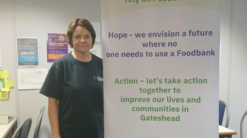 Diane Kindred standing next to a Gateshead Foodbank banner. She has dark hair and is wearing a dark blue T-shirt. The banner includes a number of action points such as "Hope - we envision a future where no one needs to use a foodbank".