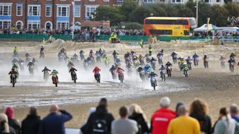 Backs of spectators with dozens of bikes coming towards them on wet sand with buildings behind