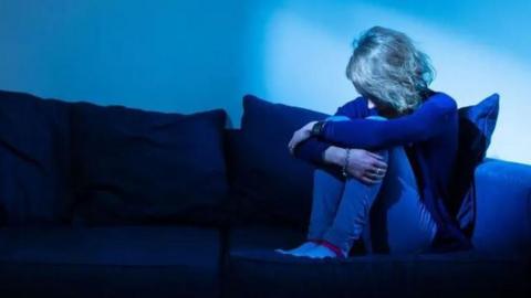 A woman sits on a sofa in a darkened room with her head down and knees pulled up to her chest. 