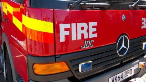 The front right-hand side of a fire engine. It is red and has fluorescent yellow checks along the side. The grill has a Mercedes Benz logo and the number plate reads WK54 BBO.