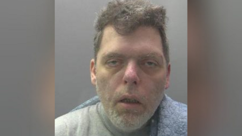 Custody photo of Karl Hutchings against a grey background. His mouth is hanging open and he has shadows under his eyes. He has some grey facial hair which is a similar colour to the hair on top of his head. 