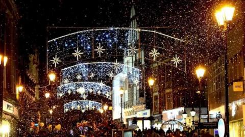 Christmas lights across Lincoln's high street at night. There are four arches of lights hanging across the street with stars on them and white lights. It's snowing and the street lamps are on.