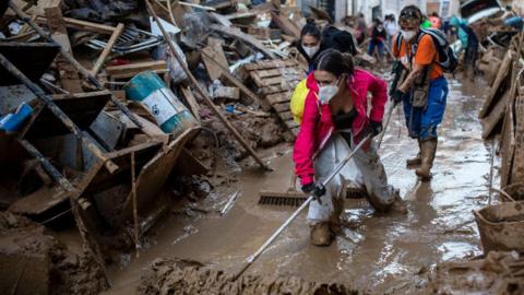 A woman, wearing a face mask, sweeps brown muddy water. There is a huge pile of muddy debris next to her. debris 