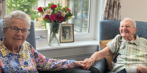 A couple holding hands in a care home. The man is smiling at the woman