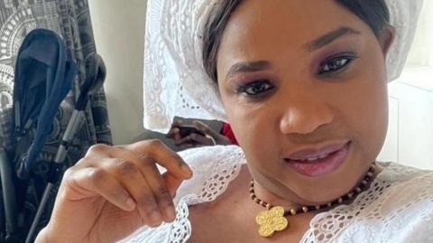 Ndata Bobb is sitting in a room in a house. She is looking into the camera and is smiling. She has a hand raised close to her face. She is wearing a white outfit decorated with embroidered circular patterns.